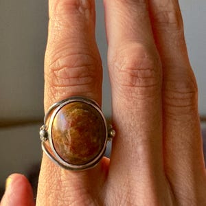 Puede incluir: Un anillo de plata con una gran piedra preciosa ovalada marrón. La piedra tiene una superficie pulida y reflectante con variaciones de color. El anillo tiene una banda decorativa de plata en espiral y pequeños acentos redondos.