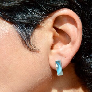 Puede incluir: Primer plano de una oreja con un pendiente rectangular plateado. El pendiente presenta un diseño azul y blanco. La persona tiene el pelo oscuro.