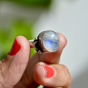 Könnte beinhalten: Ein silberner Ring mit einem großen, runden, schillernden Mondstein, der in einer Zargenfassung sitzt. Der Ring wird an einem Finger mit rotem Nagellack getragen.