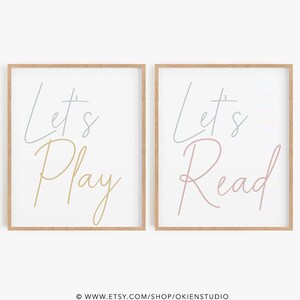 May include: Two framed prints with the text "Let's Play" in gold and "Let's Read" in pink. The prints are on a white background and have a light wood frame.