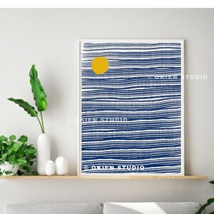 May include: Abstract art print with a yellow circle and blue and white stripes. The print is framed and hanging on a wall above a gray sofa.