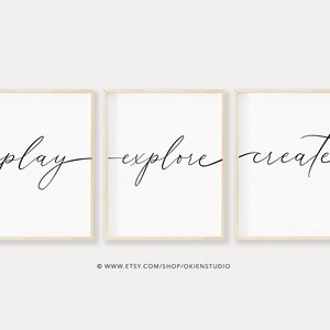 May include: Three minimalist black and white prints with the words "play", "explore", and "create" in a cursive font. The prints are framed in a light wood frame and are hung on a white wall.