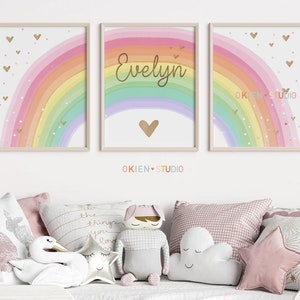 May include: Three framed prints with a rainbow design. The middle print has the name "Evelyn" written in gold script. Each print has a gold heart and gold glitter hearts scattered on a pink background.