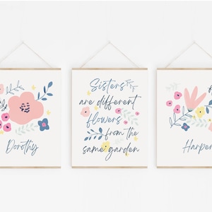 Könnte beinhalten: Drei florale Drucke mit dem Zitat "Schwestern sind verschiedene Blumen aus dem gleichen Garten". Die Drucke sind mit einem hellen Holzrahmen gerahmt und haben einen weißen Hintergrund. Die Blumen sind rosa, blau und gelb. Die Namen "Dorothy", "Harper" und das Zitat sind in Blau geschrieben.