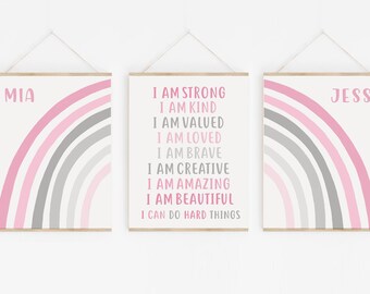 May include: Three framed prints with pink and gray rainbow designs. The middle print has the text "I am strong, I am kind, I am valued, I am loved, I am brave, I am creative, I am amazing, I am beautiful, I can do hard things". The left print has the name "MIA" and the right print has the name "JESS".