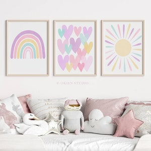 May include: Three framed prints with pastel colours. The first print features a rainbow with a yellow, orange, green, blue, and purple colour scheme. The second print features a pattern of pink, purple, and yellow hearts. The third print features a yellow sun with pastel pink, purple, and blue rays.