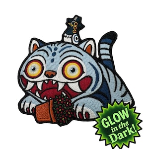 Könnte beinhalten: Gestickter Aufnäher mit einer Cartoon-Katze mit blauen Streifen, großen gelben Augen und einem roten Mund, die einen kleinen Topf mit Beeren hält. Der Aufnäher hat einen schwarzen Rand und einen grünen "GLOW in the Dark!" Stern.