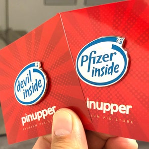 Puede incluir: Dos pines de esmalte en tarjetas rojas con lunares blancos. Los pines son plateados con texto azul. Un pin dice "devil inside" y el otro dice "Pfizer inside". Las tarjetas tienen el texto "pinupper premium pin store" en ellas.