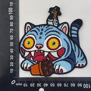 May include: Embroidered patch featuring a cartoon cat with a blue body, large yellow eyes, and a red mouth holding a flower pot. The cat has a black hat and is lying down. The patch measures approximately 4 inches in length.