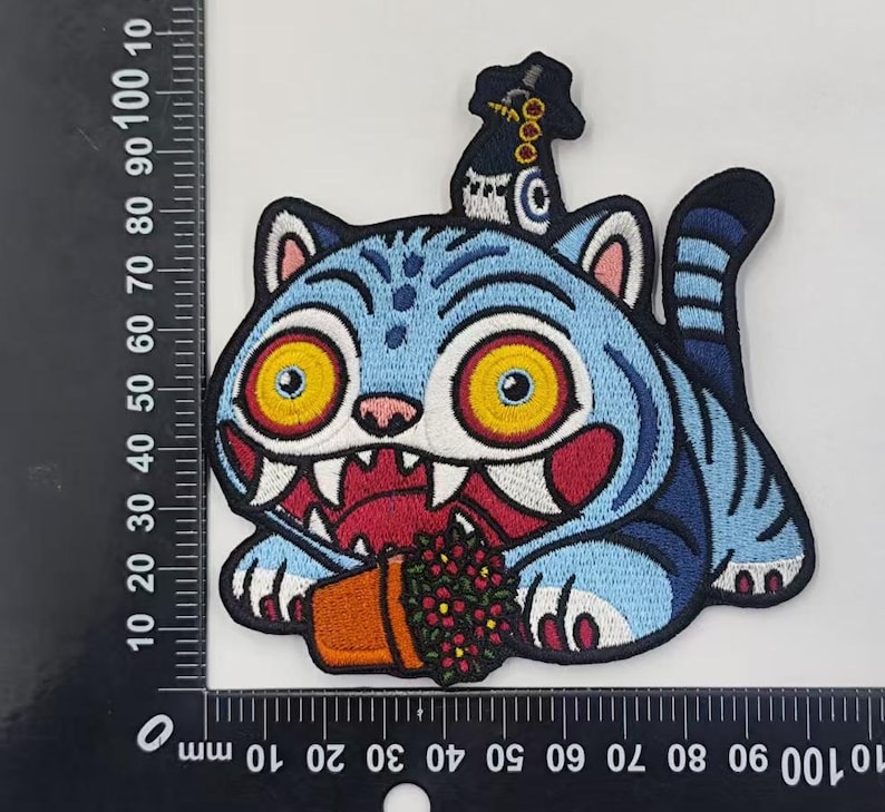 May include: Embroidered patch featuring a cartoon cat with a blue body, large yellow eyes, and a red mouth holding a flower pot. The cat has a black hat and is lying down. The patch measures approximately 4 inches in length.