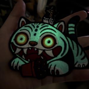 May include: A glow-in-the-dark embroidered patch featuring a cartoon tiger with large eyes, sharp teeth, and a red mouth. The tiger is holding a small pot with a plant. The patch has a black border and a small cat on top.