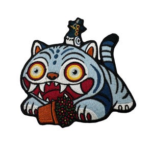 May include: Embroidered patch featuring a stylized blue cat with large yellow eyes, sharp teeth, and a red mouth. The cat is holding a small brown pot with red berries. The patch has a black border and a hat on its head.