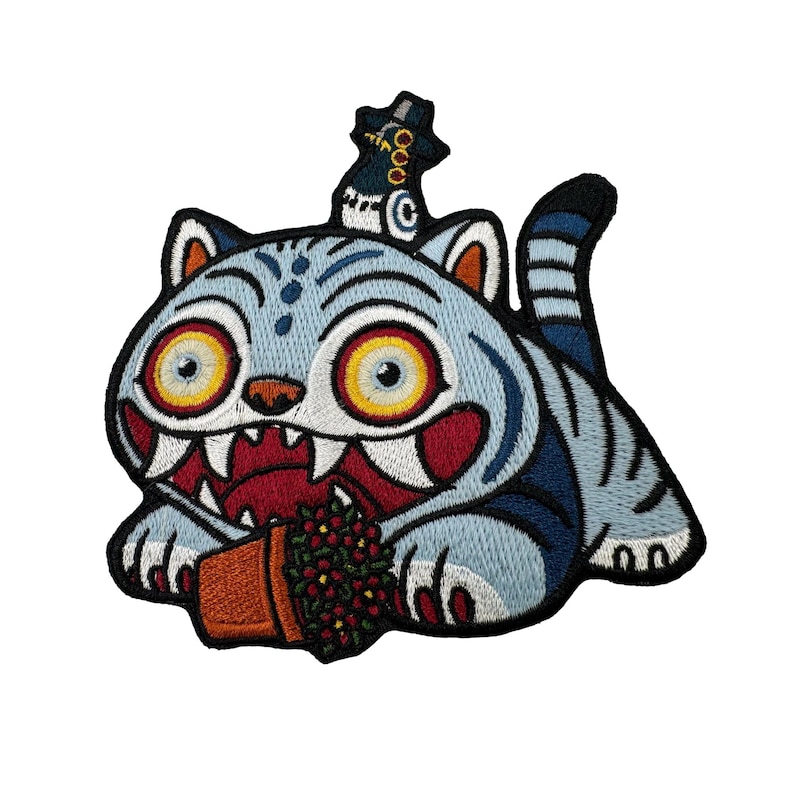 May include: Embroidered patch featuring a stylized blue cat with large yellow eyes, sharp teeth, and a red mouth. The cat is holding a small brown pot with red berries. The patch has a black border and a hat on its head.
