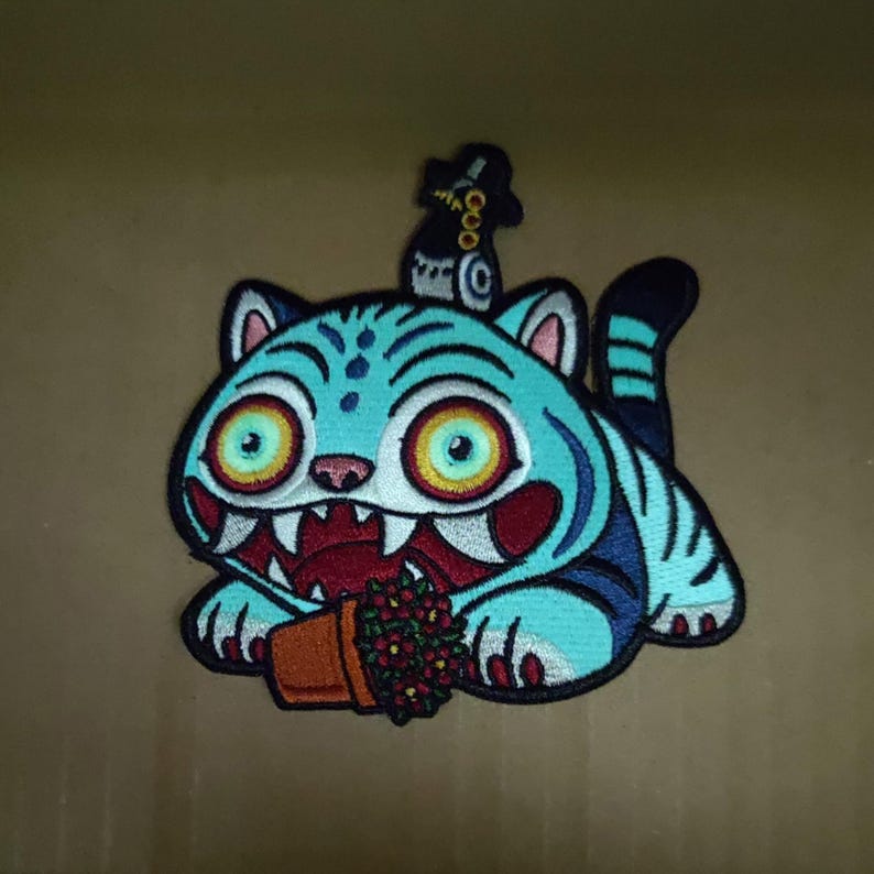 May include: Embroidered patch featuring a cartoon tiger in shades of blue, with large yellow eyes and a menacing grin. The tiger holds a small pot of red flowers. A small figure with a top hat sits on the tiger's head. The patch has a black border.