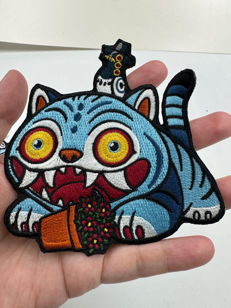 May include: Embroidered patch featuring a blue cat with yellow eyes, sharp teeth, and a flower pot. The cat has blue stripes and a black outline. A small figure sits atop the cat's head. The patch is on a white background.
