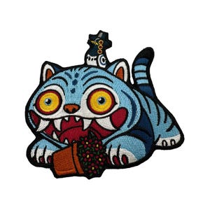 May include: Embroidered patch featuring a blue cat with yellow eyes, sharp teeth, and a small hat. The cat is holding a brown flower pot with red and green flowers. The patch has black borders and is on a white background.