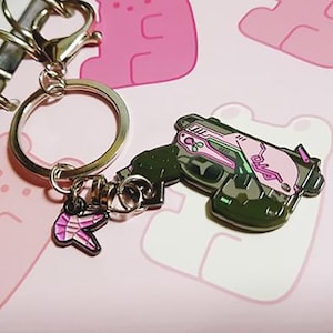 May include: A silver keychain with a pink and green gun charm and a pink and white charm. The keychain is hanging from a silver ring.