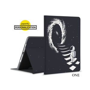 Peut inclure: Étui noir pour iPad avec un astronaute blanc montant un escalier vers une galaxie tourbillonnante. L'étui porte l'inscription "ONE" en bas.