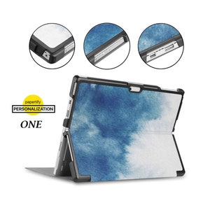 Könnte beinhalten: Eine blaue und weiße Aquarell-Hülle für ein Microsoft Surface Tablet. Die Hülle hat einen schwarzen Schutzrahmen und einen Ständer, der zum Aufstellen des Tablets ausgeklappt werden kann.
