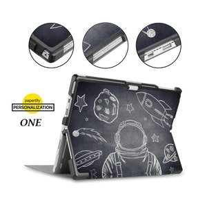 Könnte beinhalten: Schwarzes Case mit einem weißen Astronauten, einer Rakete, Sternen und Planeten Design für ein Microsoft Surface Tablet. Das Case hat einen eingebauten Ständer und eine transparente Rückseite. Das Case ist aus strapazierfähigem Kunststoff gefertigt und hat ein weiches, gummiertes Innenfutter, um Ihr Tablet vor Kratzern und Stößen zu schützen.