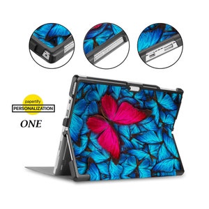 Könnte beinhalten: Eine schwarze Hülle mit einem Schmetterlingsdesign für ein Microsoft Surface Tablet. Die Hülle hat eine Standfunktion und ein blaues und pinkfarbenes Schmetterlingsmuster.