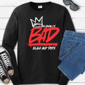 May include: Black long-sleeve sweatshirt with the text "You know I'm BAD" in red and white, and "BLACK AND DOPE" in white. The design includes a crown graphic. The image also shows black sunglasses, blue jeans, and black and white sneakers.