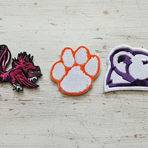 Peut inclure: Trois écussons brodés : un coq stylisé magenta et noir, une empreinte de patte orange et blanche, et une figure en forme d'ours violet et blanc. Les écussons sont sur une surface à grain de bois clair.