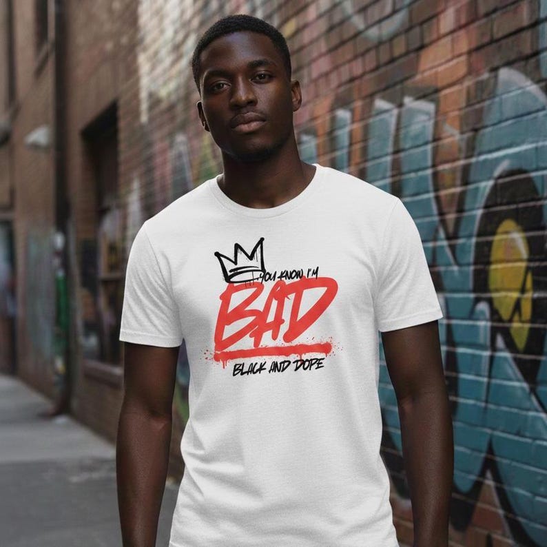 May include: White t-shirt with the text "YOU KNOW I'M BAD" in black and red, and a crown graphic. The words "BLACK AND DOPE" are below the word "BAD". The shirt is worn by a person.