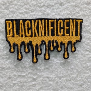 Op de afbeelding: Geborduurde patch met het woord "BLACKNIFICENT" in gele blokletters op een zwarte achtergrond. De onderkant van de patch heeft een druipend geel ontwerp met zwarte contouren, dat op gesmolten honing lijkt.