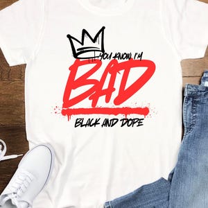 May include: White t-shirt with a black crown graphic above the red text "BAD". The phrase "YOU KNOW I'M" is above the word "BAD", and "BLACK AND DOPE" is below it. A pair of white sneakers and blue jeans are visible.
