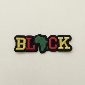 Op de afbeelding: Geborduurde patch met het woord "BLACK" in gele, rode en zwarte letters. De letter "A" is vervangen door een groene omtrek van het Afrikaanse continent. De patch heeft een zwarte rand, tegen een witte achtergrond.