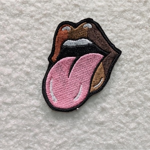 May include: Embroidered patch featuring a stylized mouth with a pink tongue sticking out. The lips are brown, with orange and tan accents, and white teeth. The patch has a black border and is likely for clothing or accessories.