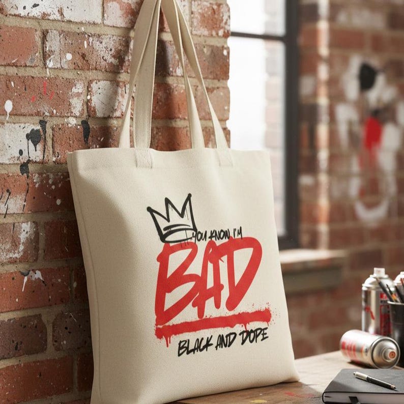 May include: Cream-colored tote bag with a black and red graphic design. The design includes a crown, the words "YOU KNOW I'M BAD" in black, and the word "BAD" in large red letters, with "BLACK AND DOPE" below.