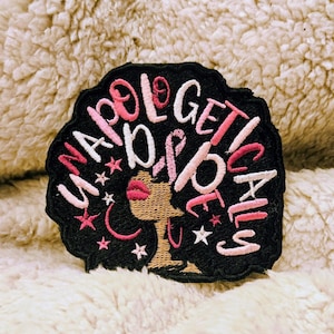 Könnte beinhalten: Schwarzer Aufnäher mit den Worten "UNAPOLOGETICALLY DOPE" in rosa und weißer Schrift. Der Aufnäher zeigt die Silhouette einer Frau mit rosa Lippen und Sternen. Der Aufnäher befindet sich auf einer flauschigen, cremefarbenen Oberfläche.
