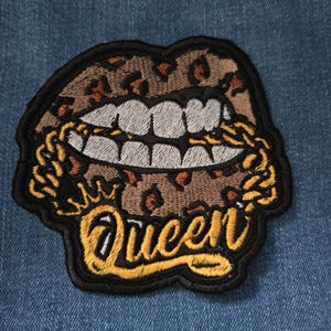 May include: A brown and black embroidered patch with a leopard print design of a mouth with gold teeth and the word "Queen" in yellow lettering.