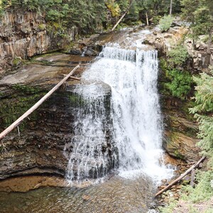 May include: A cascading waterfall flows over a rocky cliff face, surrounded by lush green trees. The water is white and foamy, creating a dramatic scene.