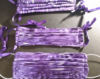 May include: Three purple face masks with white swirl patterns and purple ribbon ties. The masks are pleated and have a white elastic band.