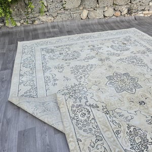 May include: A large rectangular area rug featuring a cream-colored base with intricate dark gray floral and medallion patterns. The rug has a wide decorative border and a corner is folded over to reveal the underside texture and construction.