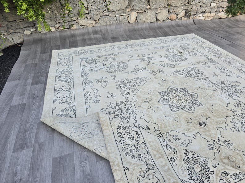 May include: A large rectangular area rug featuring a cream-colored base with intricate dark gray floral and medallion patterns. The rug has a wide decorative border and a corner is folded over to reveal the underside texture and construction.