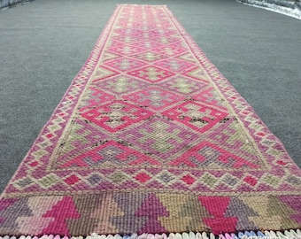 Puede incluir: Una alfombra larga y estrecha con un patrón geométrico rosa, morado y beige. La alfombra tiene flecos en ambos extremos.