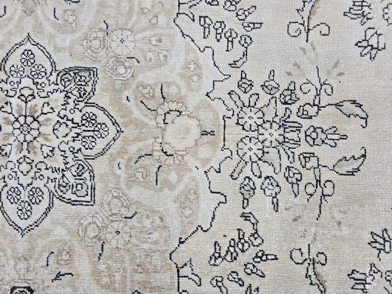 May include: A woven rug featuring a light beige background with intricate black line-drawn floral and geometric patterns. The design includes a large, detailed central medallion motif on the left side and smaller floral clusters on the right.