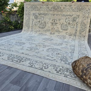 May include: A large rectangular area rug featuring a cream-colored base with intricate dark gray floral and vine patterns. The rug has a wide decorative border with repeating botanical motifs and a central medallion design, finished with fringe edges.