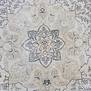 May include: A woven rug featuring a central beige floral medallion with intricate black line work. The design includes symmetrical botanical patterns and scrolling vines set against a light cream background, creating a detailed textile surface.
