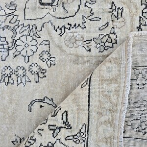 May include: A beige area rug featuring a black floral and geometric pattern. The rug is folded over at one corner, revealing the underside which displays a similar black and beige design with a visible fringe along the edge of the folded section.