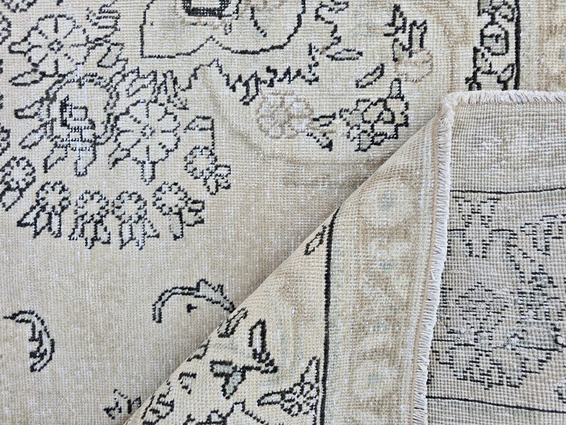 May include: A beige area rug featuring a black floral and geometric pattern. The rug is folded over at one corner, revealing the underside which displays a similar black and beige design with a visible fringe along the edge of the folded section.