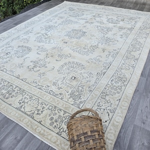 May include: A large rectangular area rug featuring a cream-colored base with intricate dark gray floral and geometric patterns. The design includes a central medallion and a detailed border. A woven brown wicker basket sits on the rug's edge.