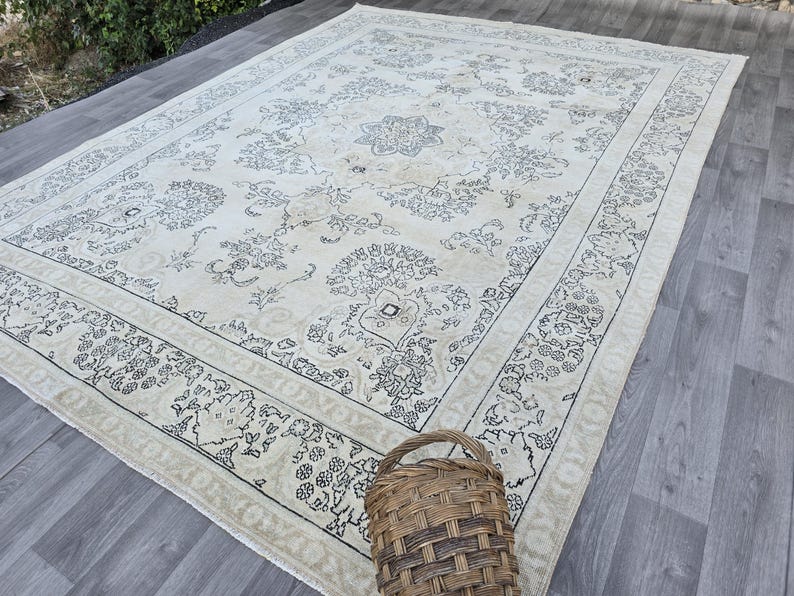May include: A large rectangular area rug featuring a cream-colored base with intricate dark gray floral and geometric patterns. The design includes a central medallion and a detailed border. A woven brown wicker basket sits on the rug's edge.