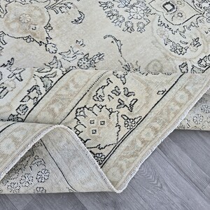 May include: A large area rug featuring a cream-colored base with intricate black and beige floral and geometric patterns. The rug is folded over to reveal the woven texture and the underside, showcasing a detailed border design with symmetrical motifs.