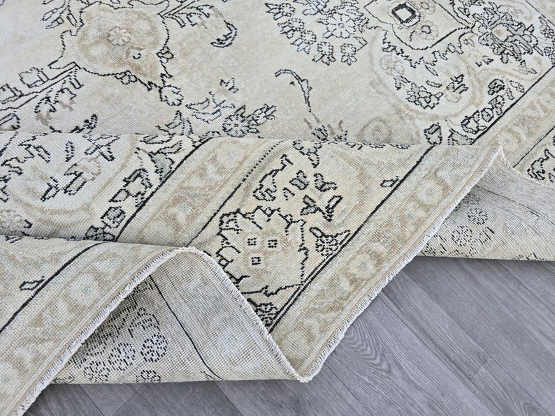 May include: A large area rug featuring a cream-colored base with intricate black and beige floral and geometric patterns. The rug is folded over to reveal the woven texture and the underside, showcasing a detailed border design with symmetrical motifs.