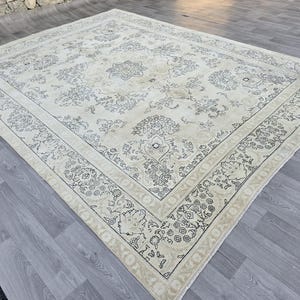 May include: A large rectangular area rug featuring a cream-colored base with intricate dark gray floral and vine patterns. The design includes a central medallion and a wide decorative border with repeating botanical motifs and a fringed edge.